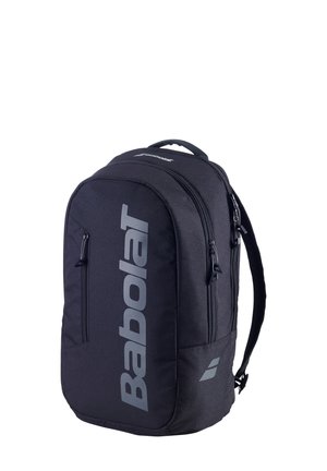 Donna con completo sportivo bianco che indossa un grande zaino nero Babolat con logo verticale, in piedi davanti a uno sfondo bianco.
