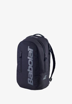 Donna con completo sportivo bianco che indossa un grande zaino nero Babolat con logo verticale, in piedi davanti a uno sfondo bianco.