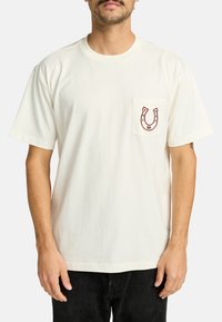 Cream cotton t-shirt featuring a horseshoe logo on the left chest pocket. Short sleeves and a classic crew neck design.