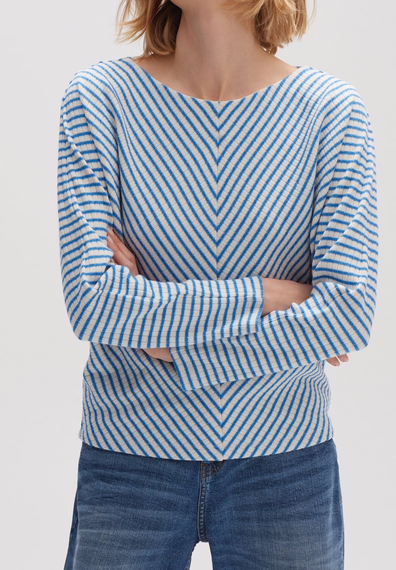 Sweter dziany z motywem jodełki w niebiesko-białe paski, szeroki dekolt, długie rękawy i prążkowany dół. Połączony z dżinsami.