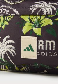 Bolsa de tela negra con un patrón floral verde. Presenta una etiqueta crema con el logo de Adidas en verde y texto. La textura parece suave y duradera.