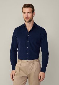 Camisa de manga larga de color azul marino, hecha de una tela con un acabado texturizado, que presenta un cuello abotonado y botones de un color contrastante.