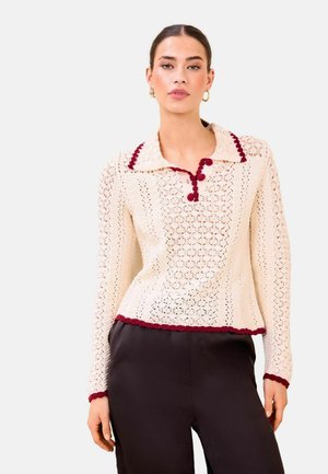 Naine, kes kannab kreemivärvi heegeldatud kampsunit burgundiapunase ääristuse ja nuppudega, kombineeritud mustade pükstega, seisab ühevärvilise tausta ees.