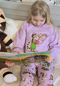 Barnet har på sig en lila långärmad skjorta med en kattgrafik och gröna detaljer, och läser en bok medan det sitter på en säng i mönstrade pyjamasbyxor.