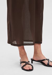 Barna áttetsző maxi ruha egyenes szegéllyel. Fekete, pántos szandálokkal viselve, alacsony sarokkal és díszes csat részlettel.