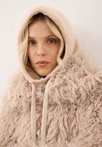 Beżowa, puszysta kurtka z kapturem wykonana z teksturowanego, kręconego materiału; posiada miękki kaptur i sznurki do regulacji. Gładka podszewka wewnętrzna kontrastuje z zewnętrzną częścią.