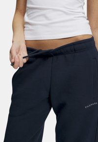 Mørkeblå joggebukser laget av mykt stoff, med elastisk liv og snor, samt sidelommer. Viser en logo i lys grå.