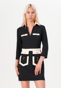 Black dress with white collar and pocket accents, featuring gold hardware, long sleeves, and a fitted design with a flared hem.