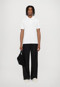 White polo shirt with short sleeves, paired with loose black trousers. Model holds a black bag, wearing white shoes. Simple and clean design.