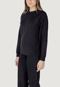 Felpa nera in cotone con maniche lunghe, colletto tondo e polsini a costine. Presenta un dettaglio logo discreto sul davanti. Abbinata a pantaloni neri.