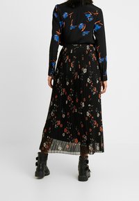 Zwarte geplisseerde rok met bloemenpatronen in rood, wit en blauw; gecombineerd met een zwarte blouse met lange mouwen met blauwe bloemenontwerpen en zwarte enkellaarsjes.