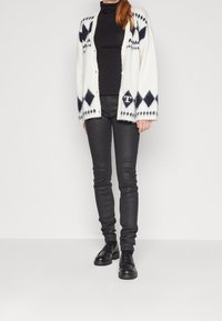 Cárdigan blanco con patrón de rombos en azul marino, cuello alto negro, jeans ajustados negros y mocasines negros. Textura suave, ajuste relajado, con botones en el frente.