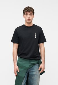 Νεαρός άνδρας με σγουρά μαλλιά που φοράει μαύρο μπλουζάκι Timberland και μπλε τζην με πράσινο φούτερ δεμένο γύρω από τη μέση του, με φόντο λευκό.