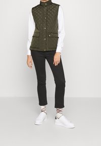 Olivgrön vadderad väst med hög krage, två framfickor och guldknappar, bärs över en vit långärmad skjorta och svarta byxor.