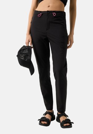 Strakke zwarte broek met roze trekkoorden, gedragen met zwarte sandalen, een cropped top en een zwarte mesh baseballpet die in één hand wordt vastgehouden.