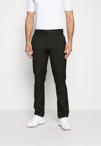Pantalons noirs avec une coupe ajustée, fabriqués dans un tissu lisse. Présentent une fermeture à bouton et un design à devant plat, portés avec une chemise blanche et des chaussures blanches.
