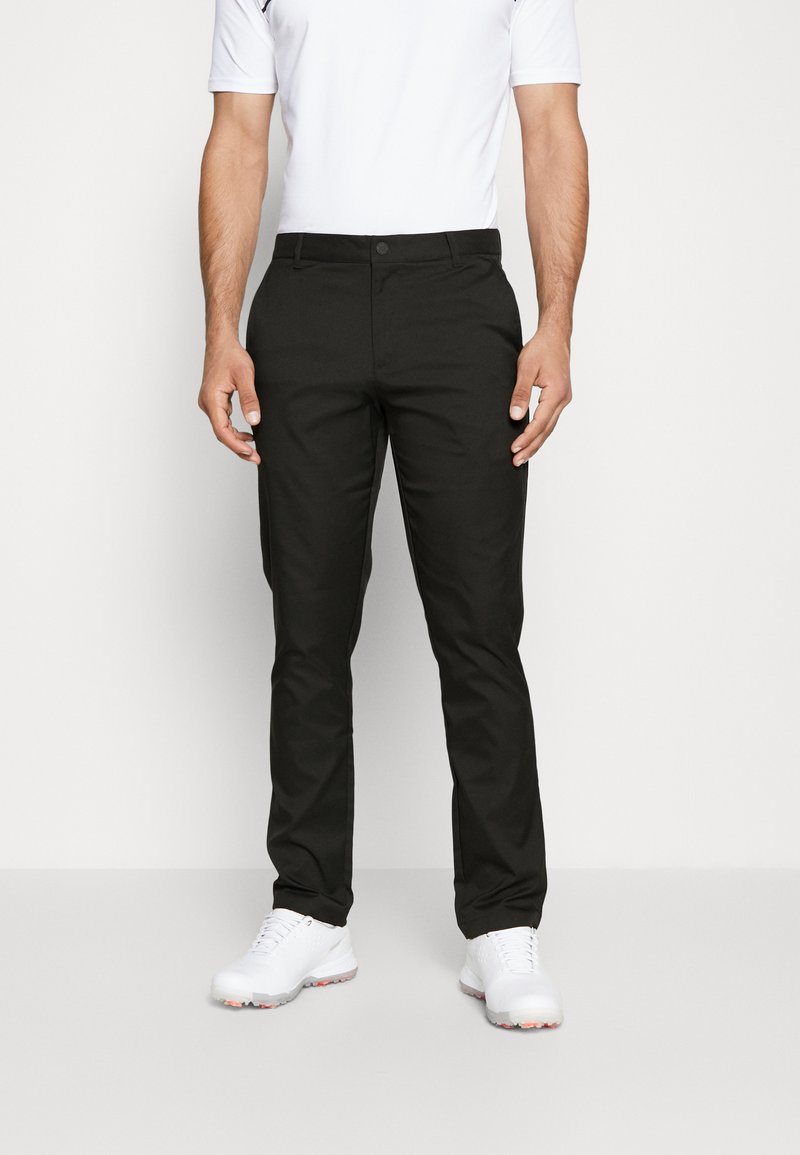Pantalons noirs avec une coupe ajustée, fabriqués dans un tissu lisse. Présentent une fermeture à bouton et un design à devant plat, portés avec une chemise blanche et des chaussures blanches.
