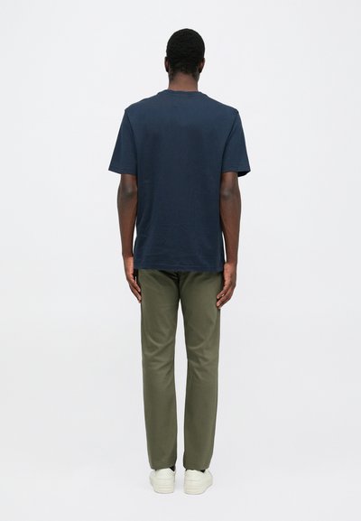 Mann in einem navyblauen Kurzarmhemd, olivgrünen Hosen und weißen Sneakern, der mit dem Gesicht zur Wand steht und einen schlichten Hintergrund hat.