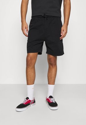 Homme portant un short noir à cordon, des chaussettes blanches à tige mi-haute et des baskets basses noires et rouges, debout devant un fond blanc uni.