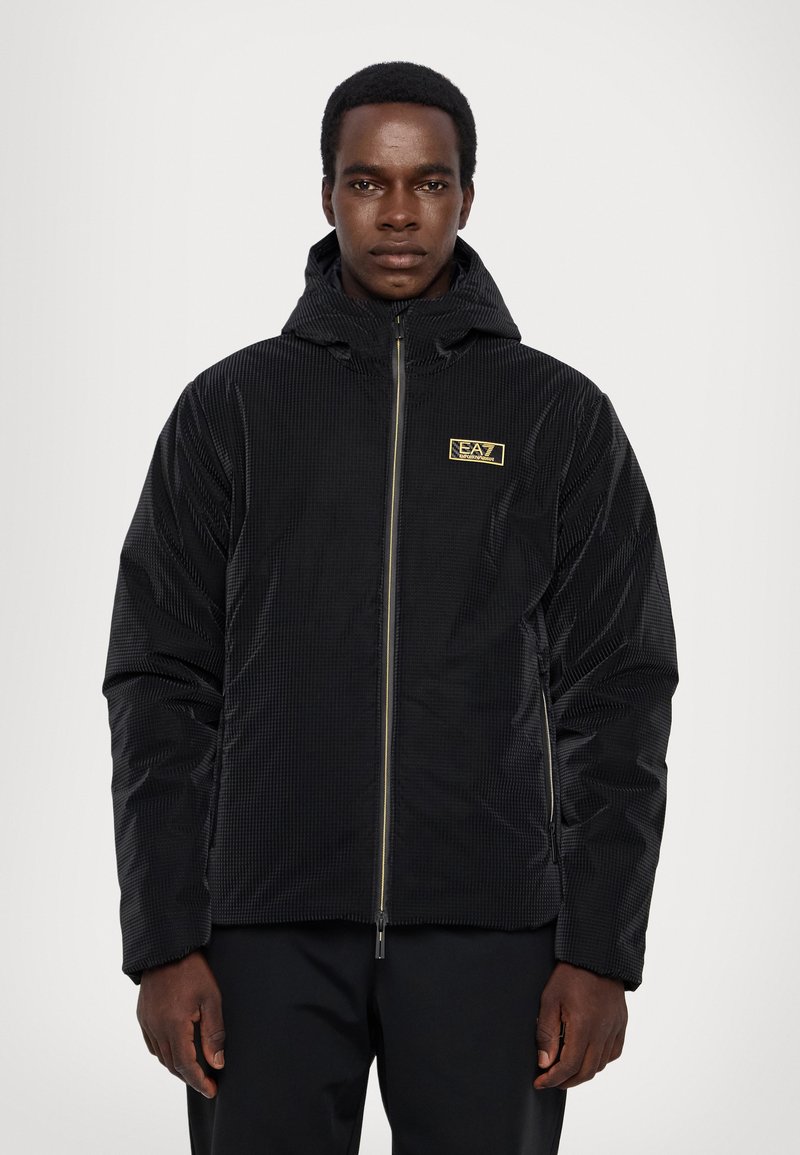 Giacca nera con zip e cappuccio, caratterizzata da un motivo testurizzato e una piccola etichetta con logo dorato sul petto. Tessuto morbido e leggero.