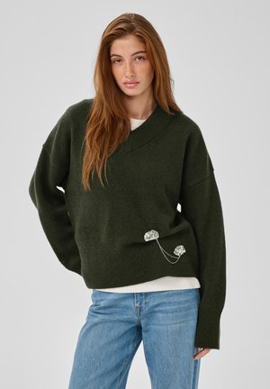 Ung kvinde med langt rødt hår iført en mørkegrøn oversized sweater, lyseblå jeans og en hvid skjorte under, stående med hånden på hoften.