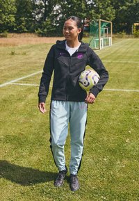 Melna zip-up džeka ar purpura akcentiem un logo, gaiši zili sporta bikses ar melnām sānu līnijām, turot rakstainu futbola bumbu uz zāli.