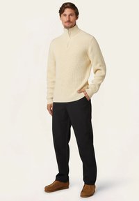 Beiger gerippter Pullover mit hohem Kragen und Viertel-Reißverschluss, kombiniert mit schwarzen Hosen und braunen Slippern, getragen von einem stehenden Mann.