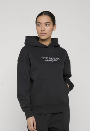 Femme portant un sweat à capuche noir Rocawear avec poche avant, regardant légèrement sur le côté sur un fond clair uni.