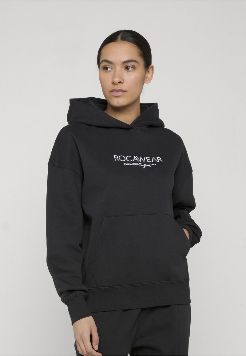 Γυναίκα με μαύρο φούτερ Rocawear με εμπρόσθια τσέπη, κοιτάζει ελαφρώς προς τα πλάγια σε απλό φωτεινό φόντο.