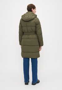 Oliven grøn pufferjakke med hætte, bælte i taljen og quiltet tekstur, parret med blå vidde jeans og sorte sko.