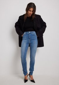 Vaqueros azules de talle alto en un corte slim, hechos de denim con una textura suave. Combinados con un suéter negro de canalé y un abrigo negro texturizado.