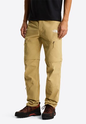 Hombre con pantalones de senderismo convertibles beige con piernas desmontables mediante cremallera y zapatos de senderismo marrones, de pie contra un fondo blanco.