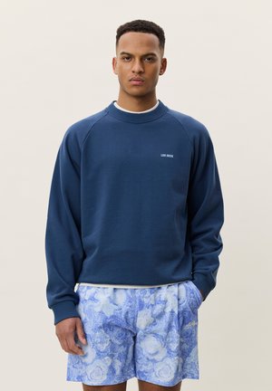 Jeune homme portant un sweat bleu marine avec un petit texte blanc et un short bleu à motifs, debout avec une main dans la poche.