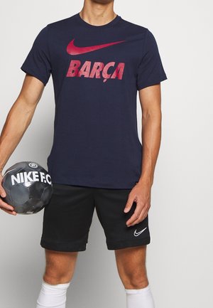 Granatowa koszulka Nike z dużym czerwonym logo "BARÇA". Połączona z czarnymi szortami i trzymana w rękach czarno-biała piłka nożna Nike F.C.