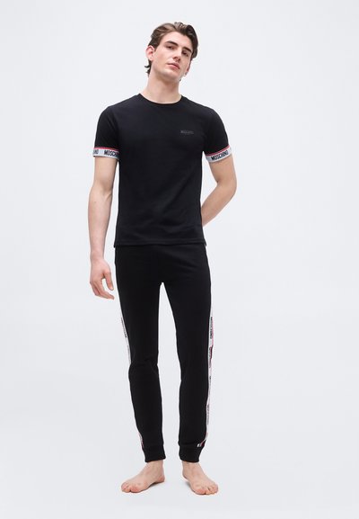 Jeune homme debout, pieds nus, portant un t-shirt noir Moschino et un pantalon de survêtement noir assorti avec des bandes à logo sur les manches et les côtés du pantalon.