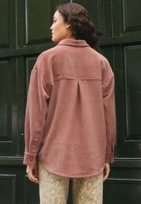 Camicia rosa a maniche lunghe, con trama e polsino con bottoni, e piega posteriore. Indossata sopra pantaloni beige con un sottile motivo leopardato.