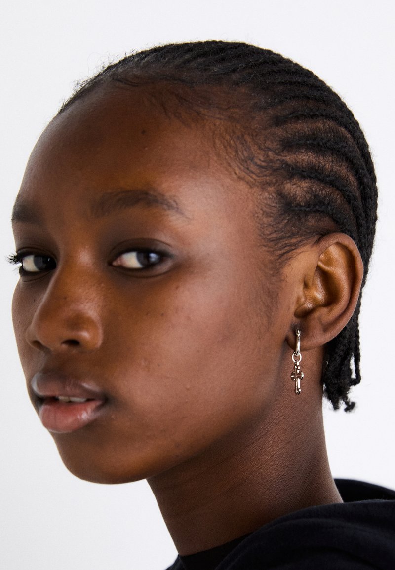 Ασημί σκουλαρίκι με κρίκο και κρεμαστό σταυρό, φορεμένο από άτομο με πλεξούδες. Ο τόνος του δέρματος είναι σκούρος και το φόντο είναι λευκό.