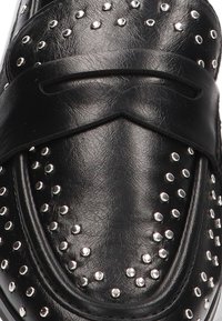 Mocasines de cuero negro con una superficie texturizada, adornados con tachuelas de plata y un diseño de tiras entrecruzadas en la parte superior. Acabado suave.