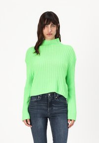 Pulover verde striat, strălucitor, cu guler înalt și manșete strâmte, purtat cu blugi negri. Puloverul are o croială lejeră și scurtă.