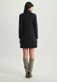 Robe noire sans manches avec un col haut, présentant un corsage ajusté et une jupe évasée. Accompagnée de bottes hautes en cuir à imprimé serpent.