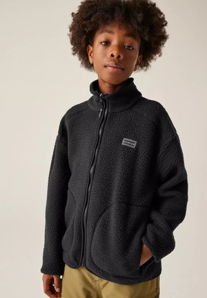 Jong persoon met krullend haar, draag een zwarte fleece met ritssluiting met het logo "DARE 2B" en beige broek, staand met één hand in de zak.