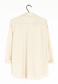 Vaaleanbeige pitkähihainen paita, jossa on kaulus ja joka on valmistettu teksturoidusta kankaasta. Ominaisuuksiin kuuluu väljä istuvuus ja pyöristetty helma käännetyillä hihansuilla.