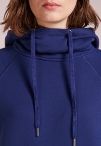Felpa con cappuccio blu navy con colletto alto e ripiegato, dotata di lacci con punte metalliche e maniche raglan, realizzata in tessuto morbido.