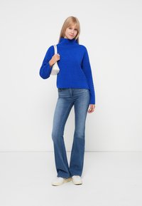 Un pull col roulé en laine bleu avec une coupe ample, associé à un jean bleu clair évasé et des baskets blanches, tenant un petit sac à main blanc.