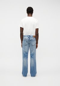 Άτομο που φοράει λευκό κοντομάνικο μπλουζάκι και φαρδιά ανοιχτό μπλε τζιν, στέκεται αντικρίζοντας ένα απλό λευκό φόντο.