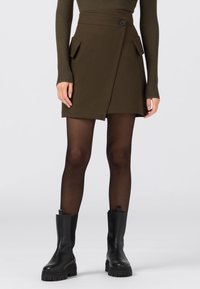 Olivgrön omslagskjol med framfickor, enkel knappstängning och texturerad tyg. Bärs med svarta ankelboots och genomskinliga strumpbyxor.