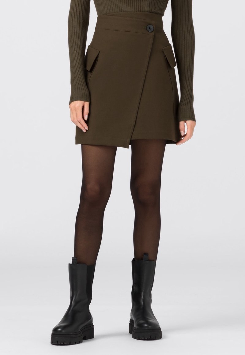 Olivgrön omslagskjol med framfickor, enkel knappstängning och texturerad tyg. Bärs med svarta ankelboots och genomskinliga strumpbyxor.