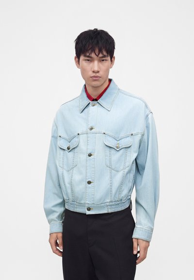 Jeune homme aux cheveux noirs courts portant une veste en jean bleu clair sur une chemise rouge et un pantalon noir, debout devant un fond blanc uni.