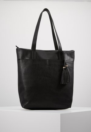 Zwarte leren tote tas met dubbele lange banden en twee decoratieve kwastjes aan één kant, geplaatst op een wit voetstuk tegen een effen achtergrond.