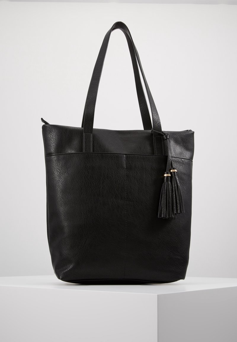 Sac cabas en cuir noir avec deux longues anses et deux pompons décoratifs suspendus d'un côté, posé sur un piédestal blanc sur un fond uni.